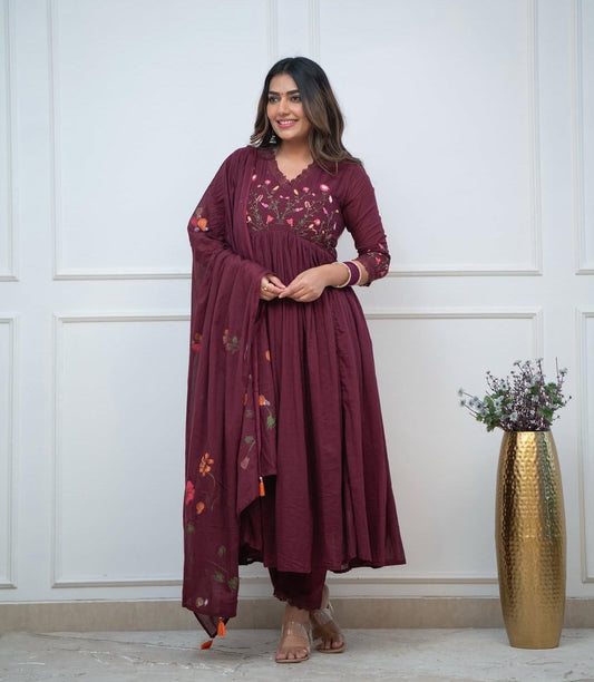 Woman wearing a maroon traditional outfit with floral embroidery, standing against a white paneled wall.